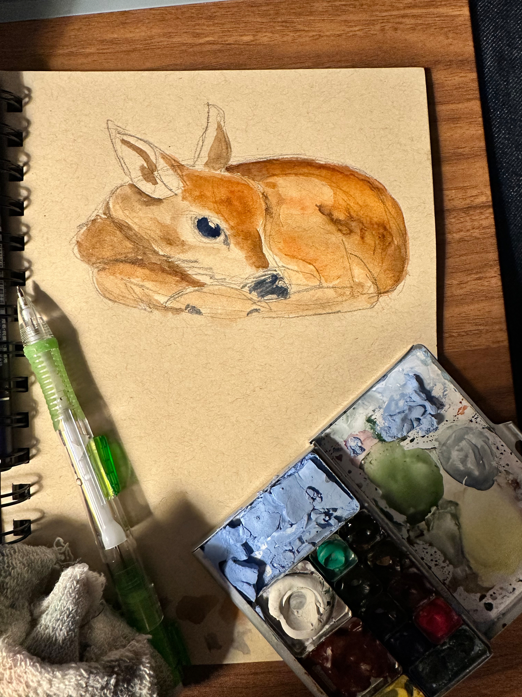 A watercolor painting of a resting deer is depicted on a sketchbook, surrounded by a paint palette, a mechanical pencil, and a cloth.
