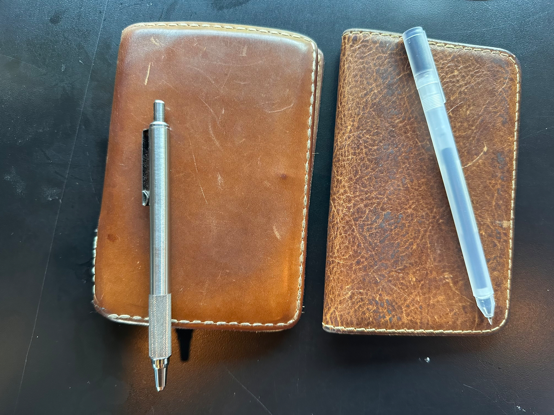 Two brown leather notebooks are placed on a black surface, each with a pen resting on top.