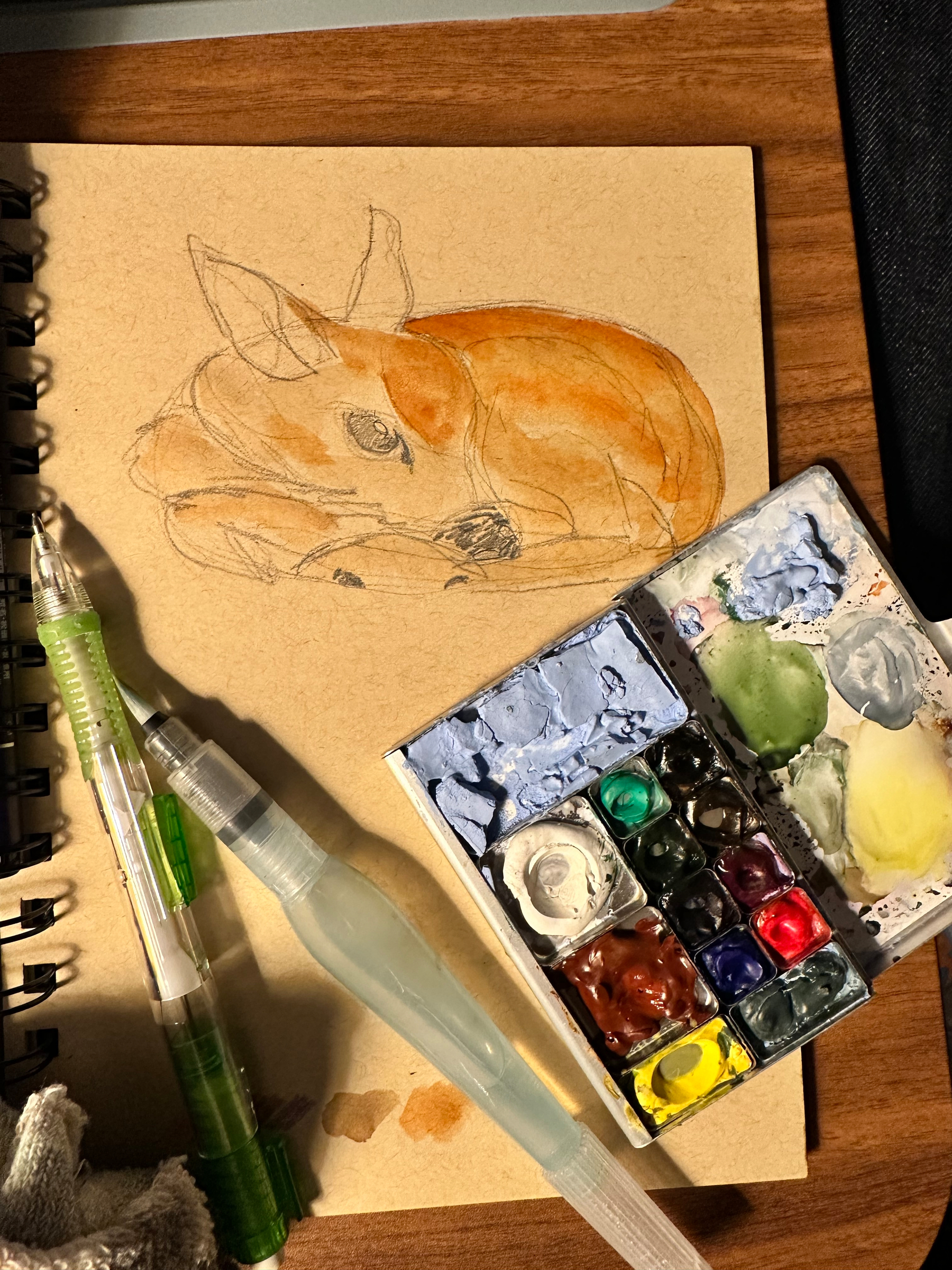 A watercolor sketch of a resting dog is displayed alongside paintbrushes and a colorful watercolor palette on a wooden surface.