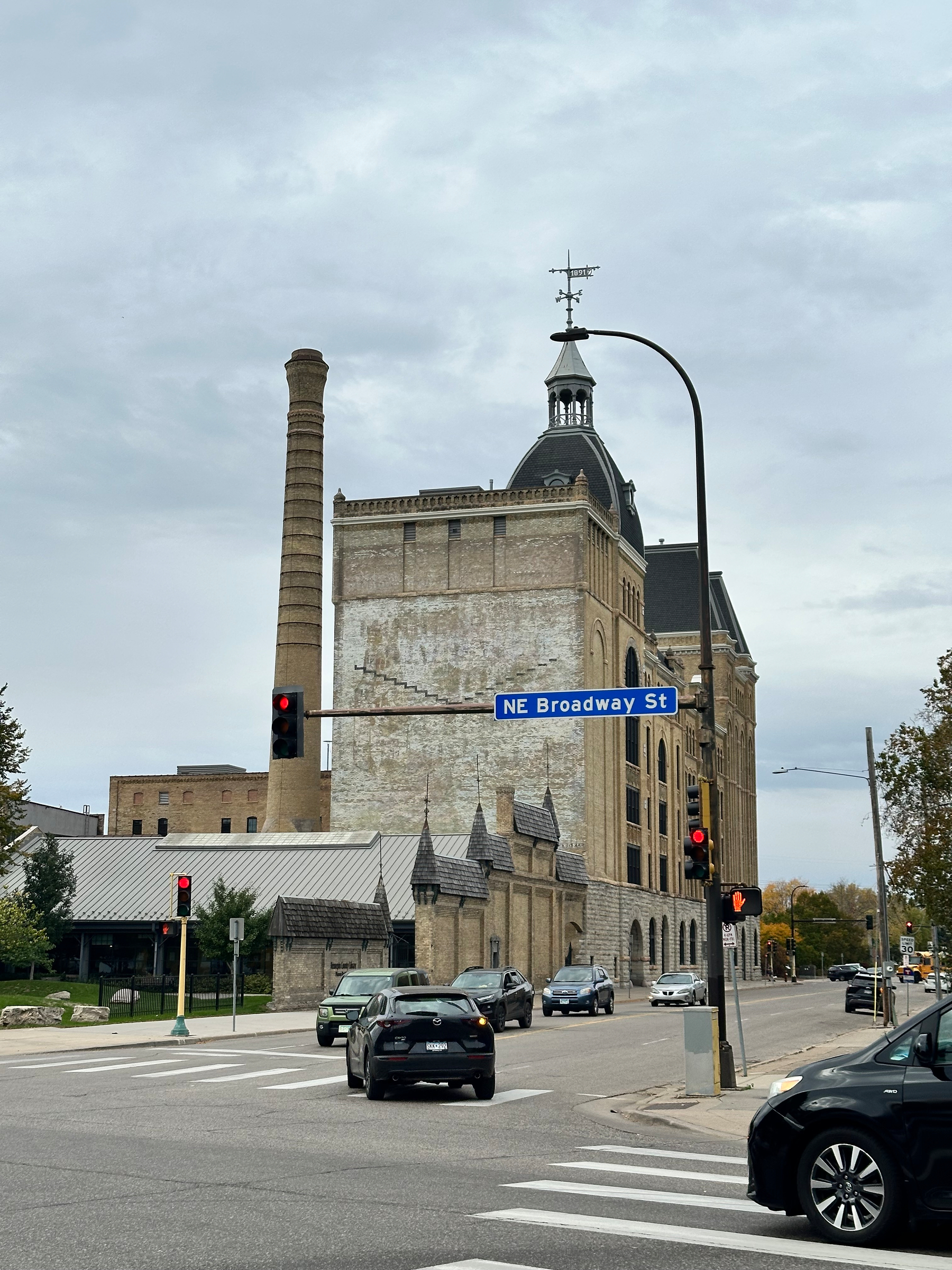 A historic brick building with a tall smokestack stands at the corner of a street intersection marked by traffic lights and a NE Broadway St sign under a cloudy sky.