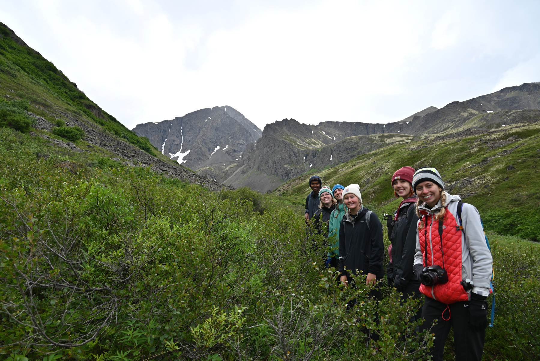 A group of seven people dressed in outdoor clothing stand on a green hillside with mountains in the background under a cloudy sky.