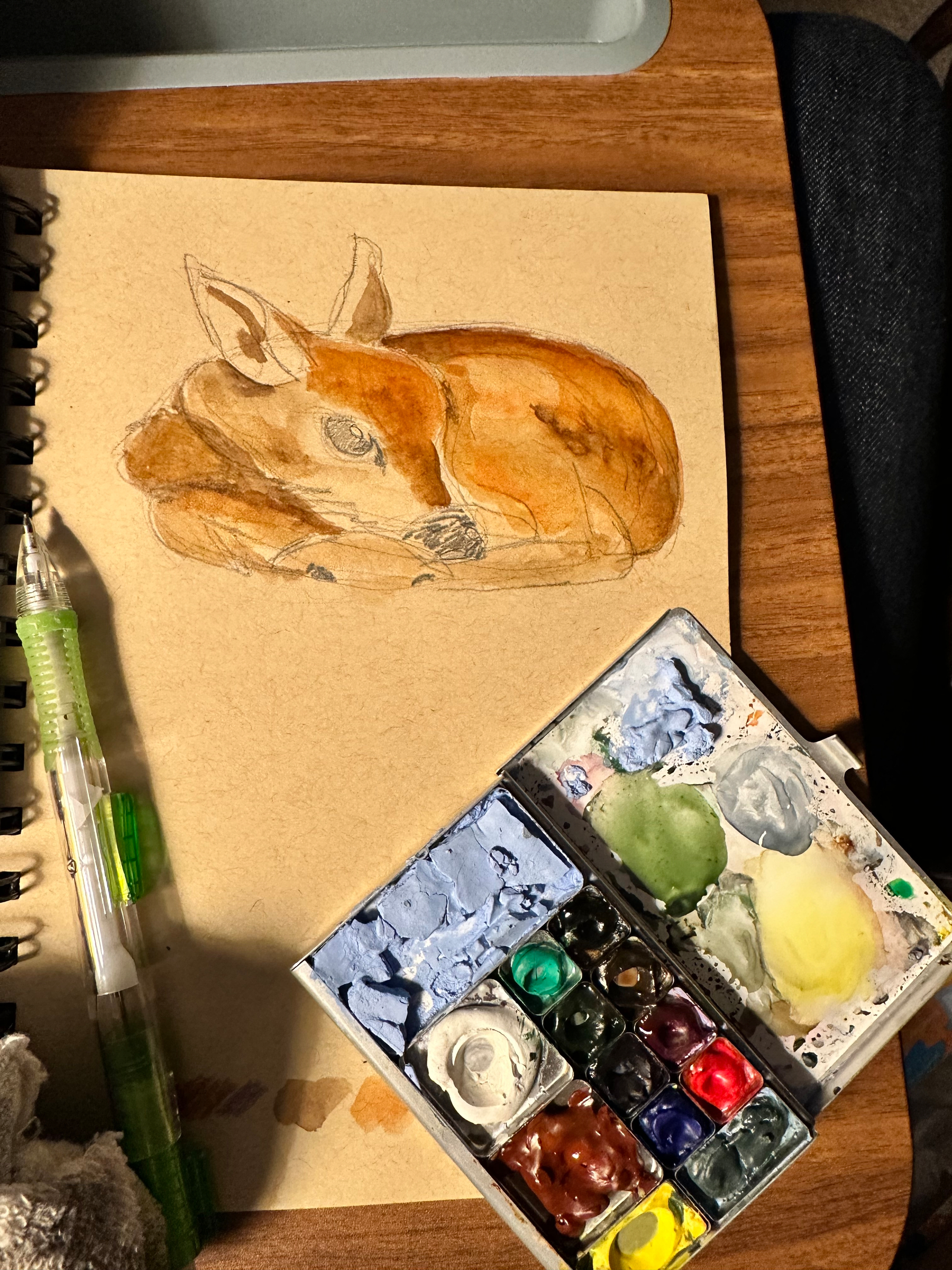 A watercolor painting of a resting fawn is shown alongside a palette of paints and a pen on a wooden surface.
